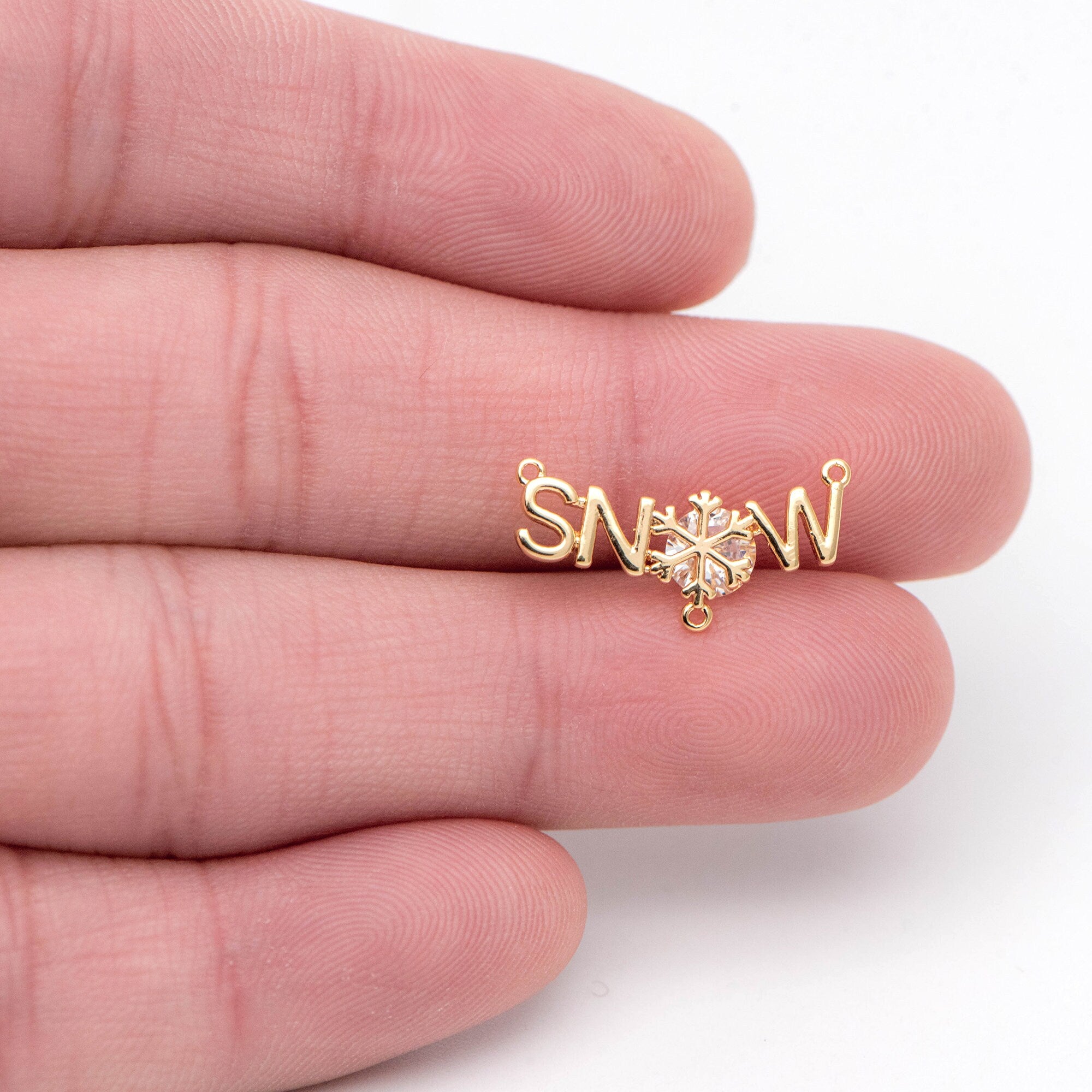 May include: A gold charm with the word 'SNOW' spelled out in cursive letters. A snowflake with a clear rhinestone center is in the center of the charm. The charm has two jump rings for attaching to a necklace or bracelet.