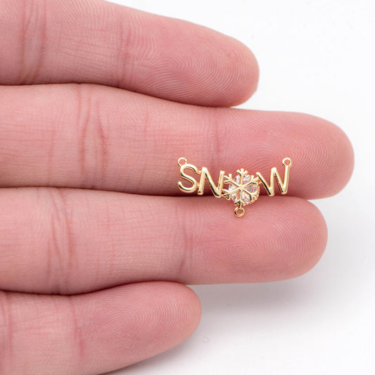 May include: A gold charm with the word 'SNOW' spelled out in cursive letters. A snowflake with a clear rhinestone center is in the center of the charm. The charm has two jump rings for attaching to a necklace or bracelet.