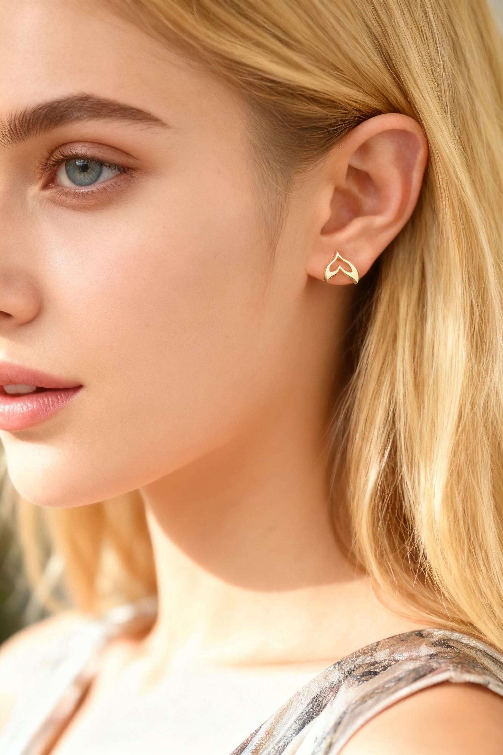 May include: A close-up of a person's ear with a gold-colored earring. The earring has a stylized, open design, resembling a flame or a stylized heart. The person has fair skin and blonde hair.