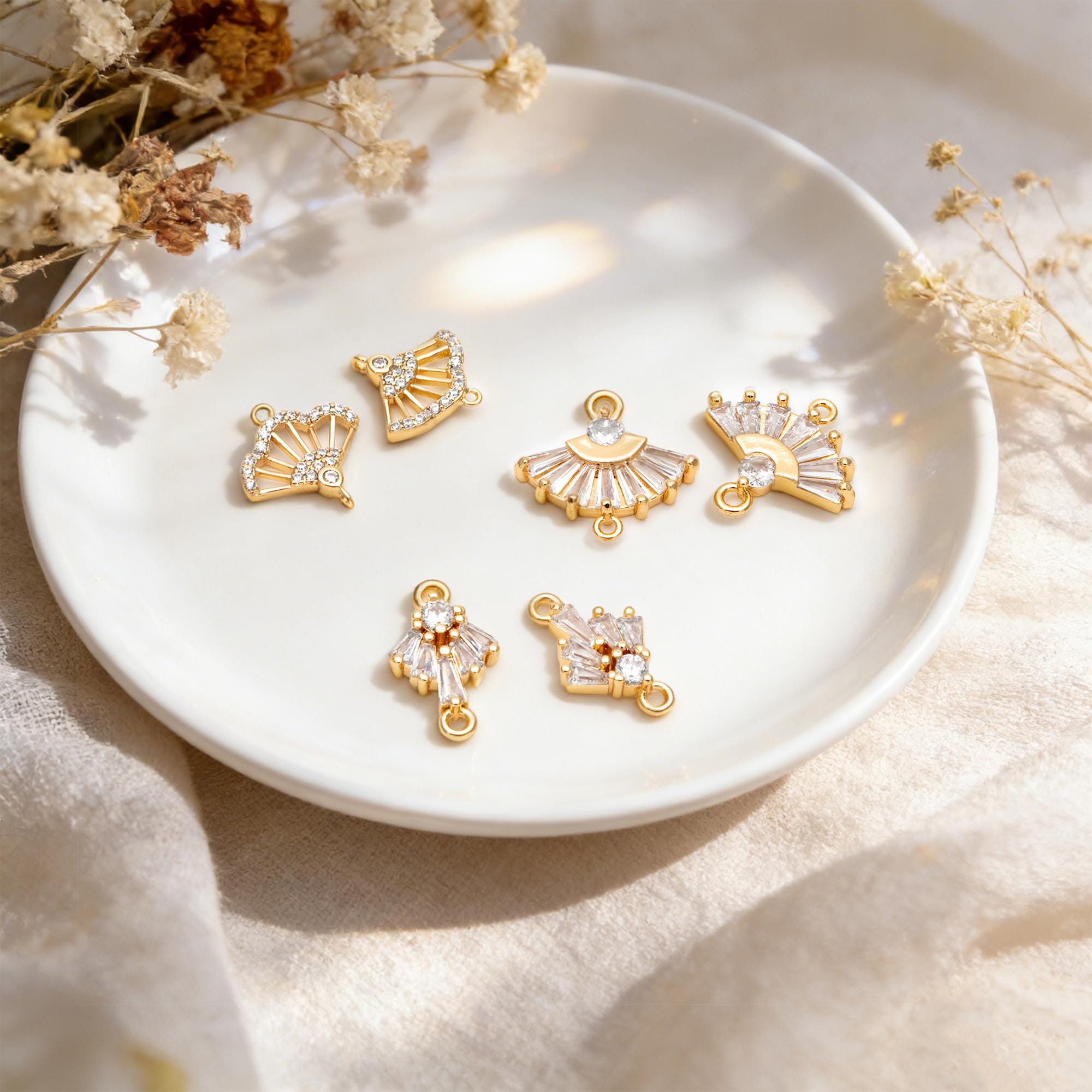 May include: Gold-toned jewelry charms in various fan and geometric shapes, adorned with clear crystals. The charms are displayed on a white plate, with dried flowers in the background. These charms are ideal for jewelry making.