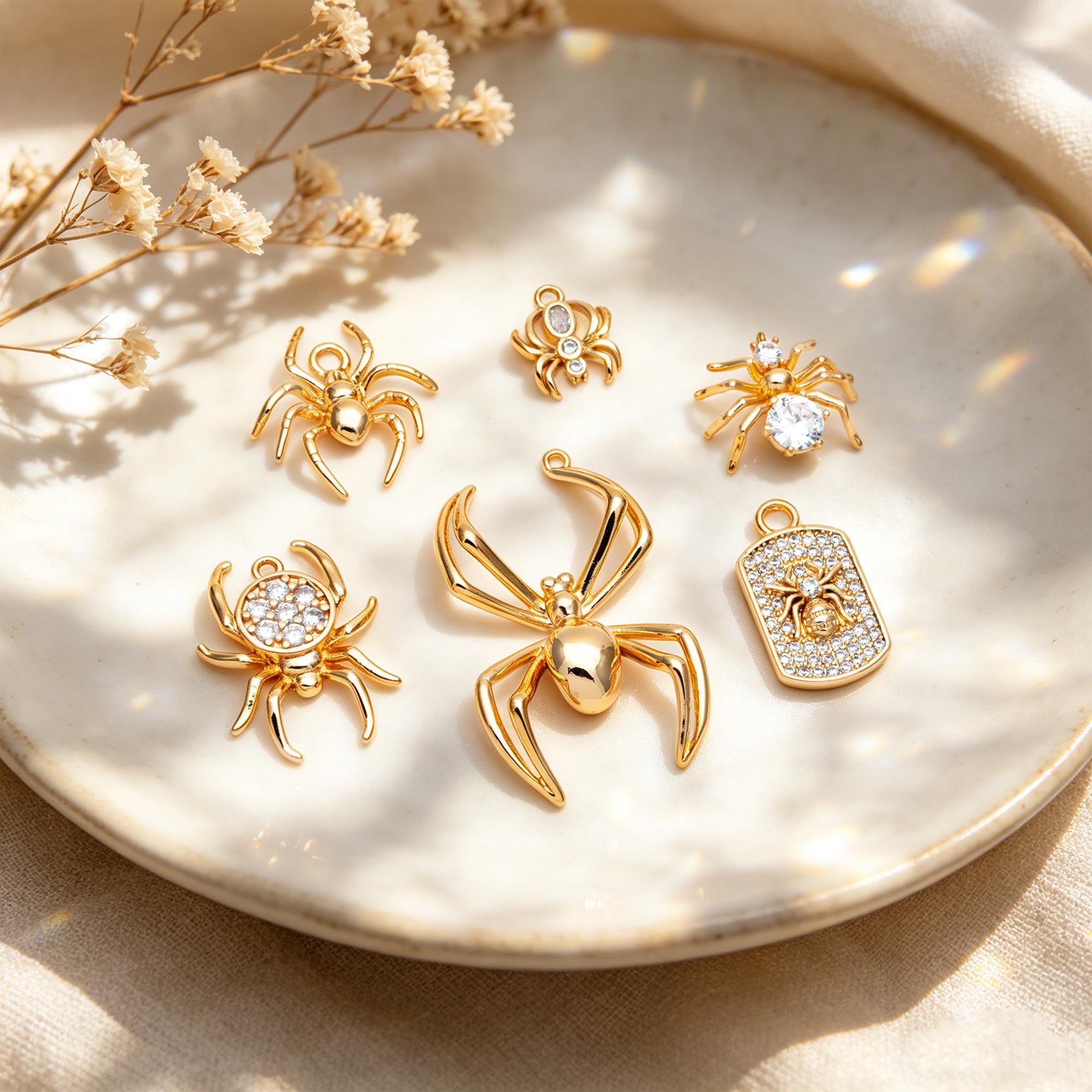 May include: A collection of gold-colored spider charms, some adorned with clear crystals. The charms vary in size and design, including a rectangular pendant with a spider motif. The charms are displayed on a white plate.