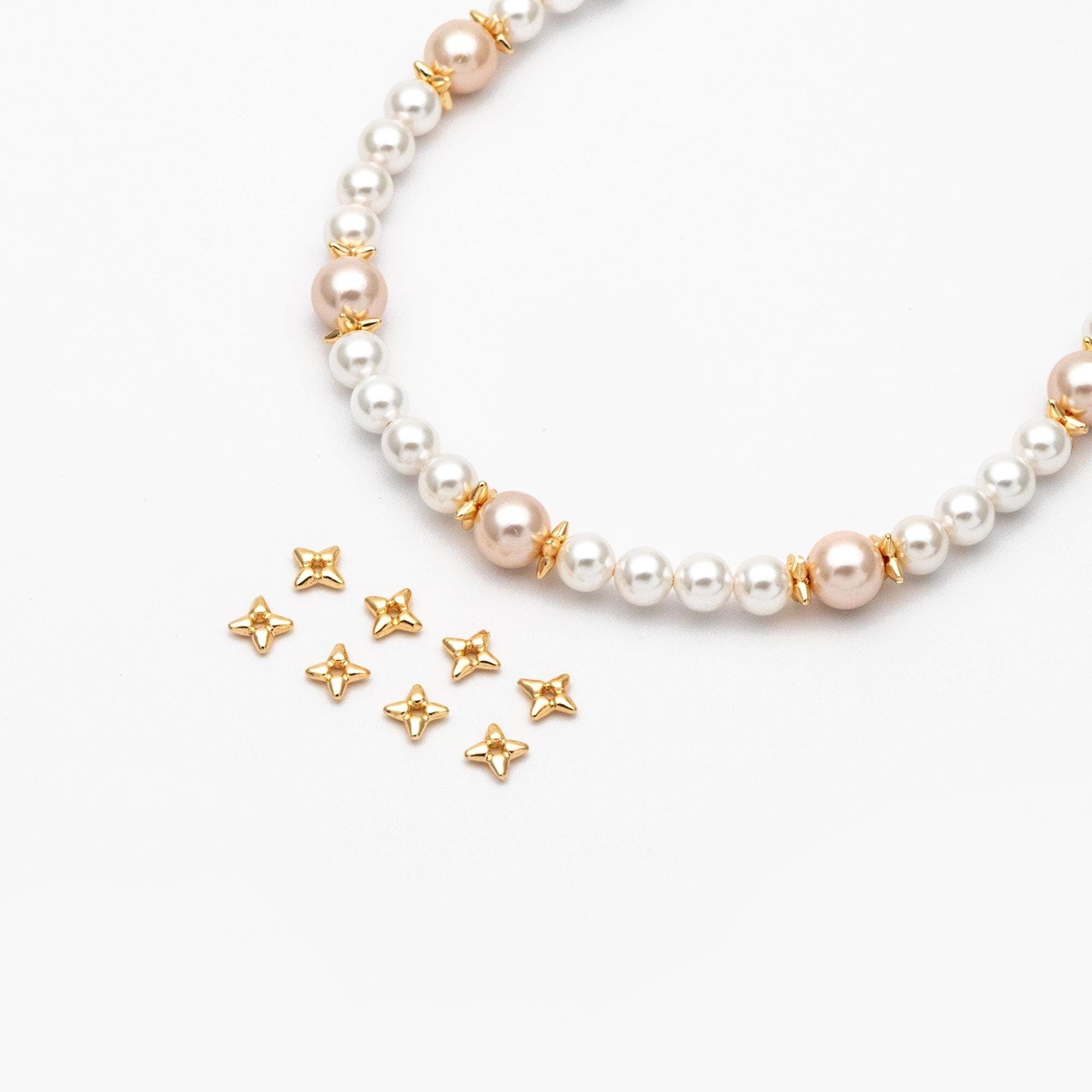 May include: A white and pink pearl necklace with gold star-shaped beads. The necklace is laid out on a white surface with additional gold star-shaped beads scattered around it.