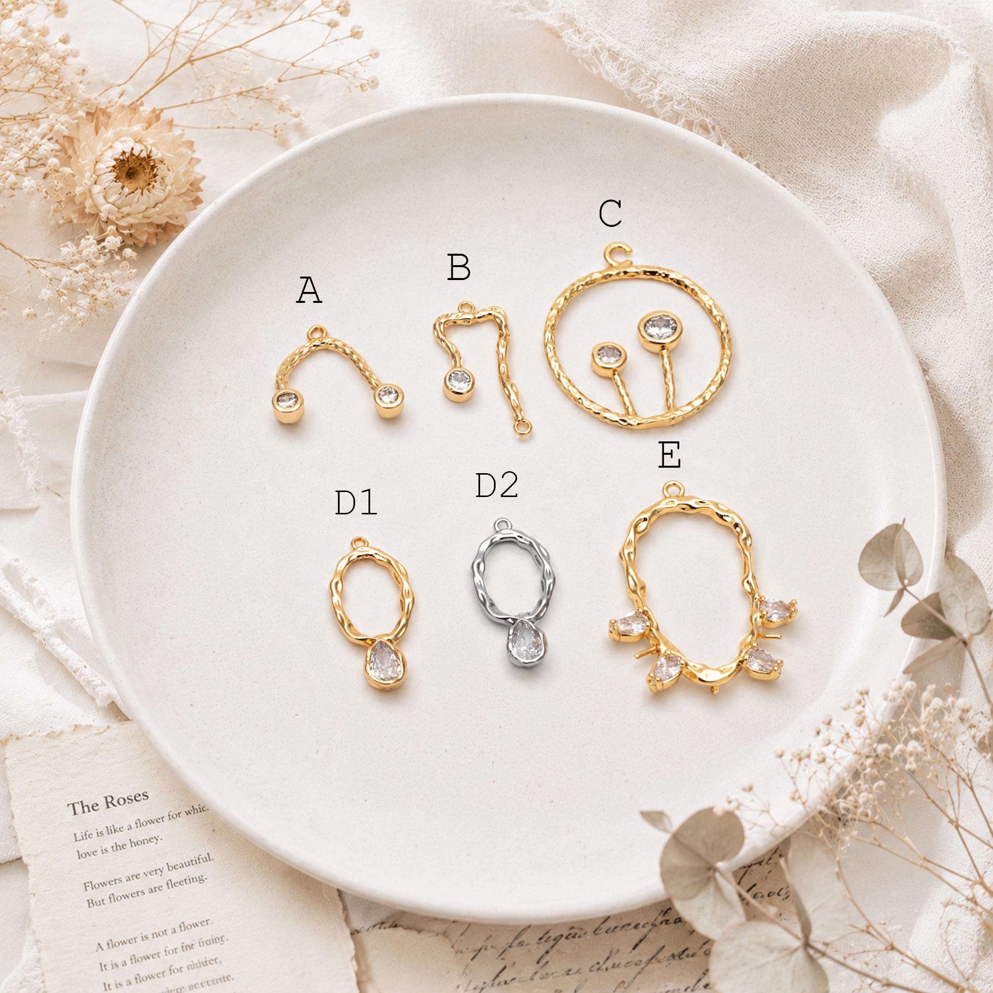May include: A collection of metal jewelry charms labeled A, B, C, D1, D2, and E. Charms A, B, C, and E are gold-toned with clear stones. Charm D1 is gold-toned with a clear stone, and D2 is silver-toned with a clear stone. Each piece has a loop.