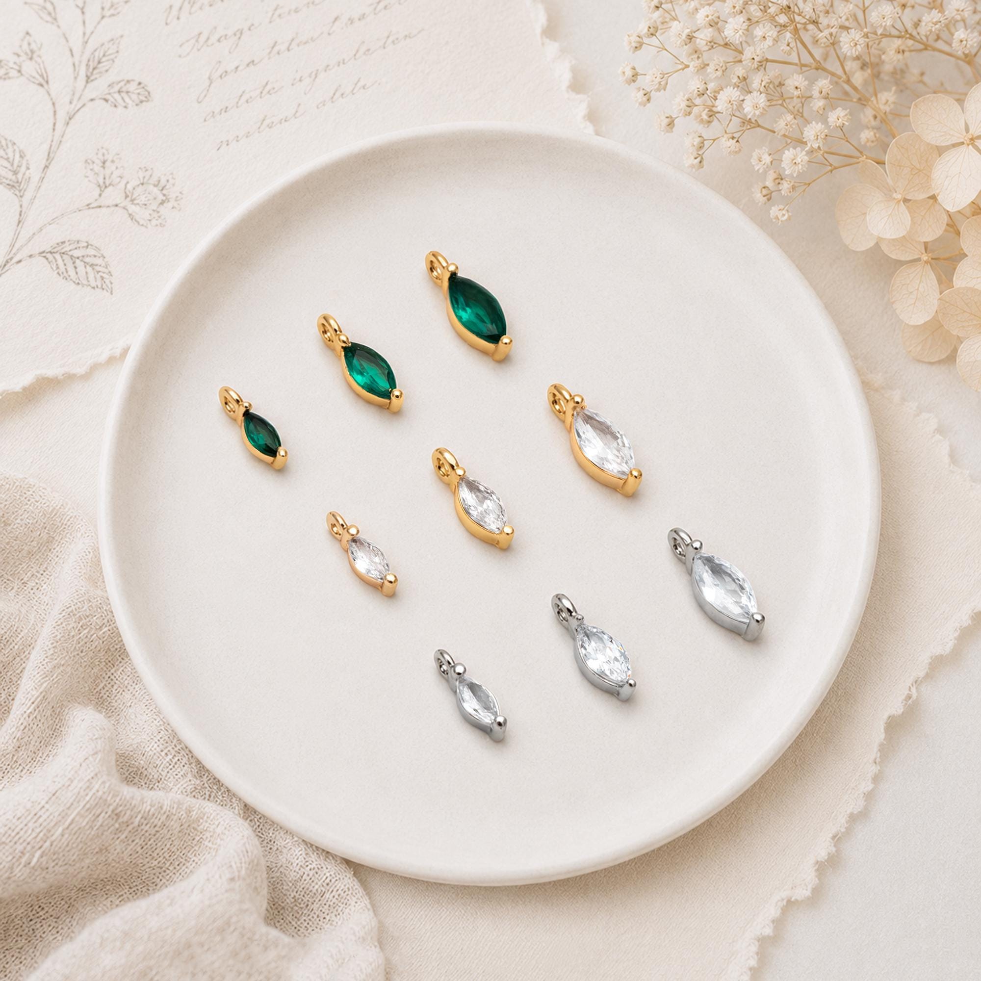 May include: A collection of marquise-cut jewelry charms in various sizes and colors. The set includes gold-toned charms with emerald green and clear stones, alongside silver-toned charms with clear stones, each featuring a small top loop for attachment.