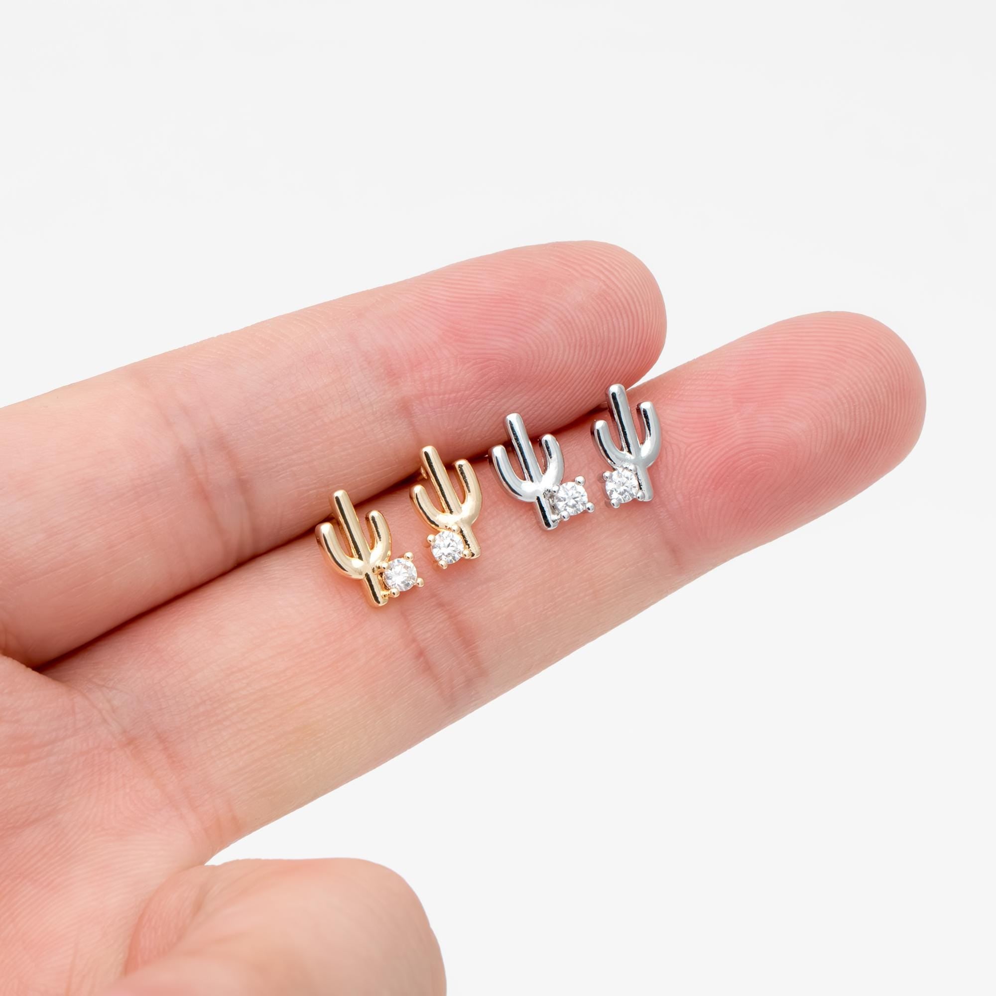 May include: Four cactus-shaped stud earrings. Two are gold-toned, and two are silver-toned. Each earring features a small, clear gemstone at the base. The earrings are displayed on a light background.