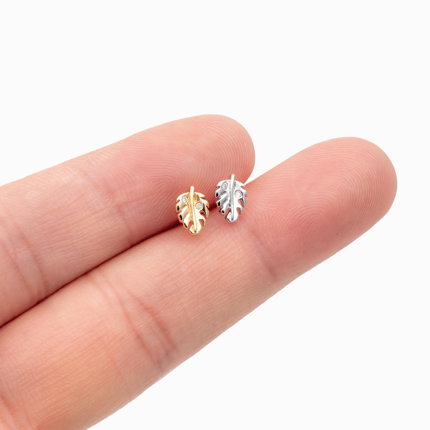 May include: Two small leaf-shaped earrings, one gold-toned and one silver-toned, are displayed. The gold earring has three small clear stones. The earrings are held between two fingers against a white background.