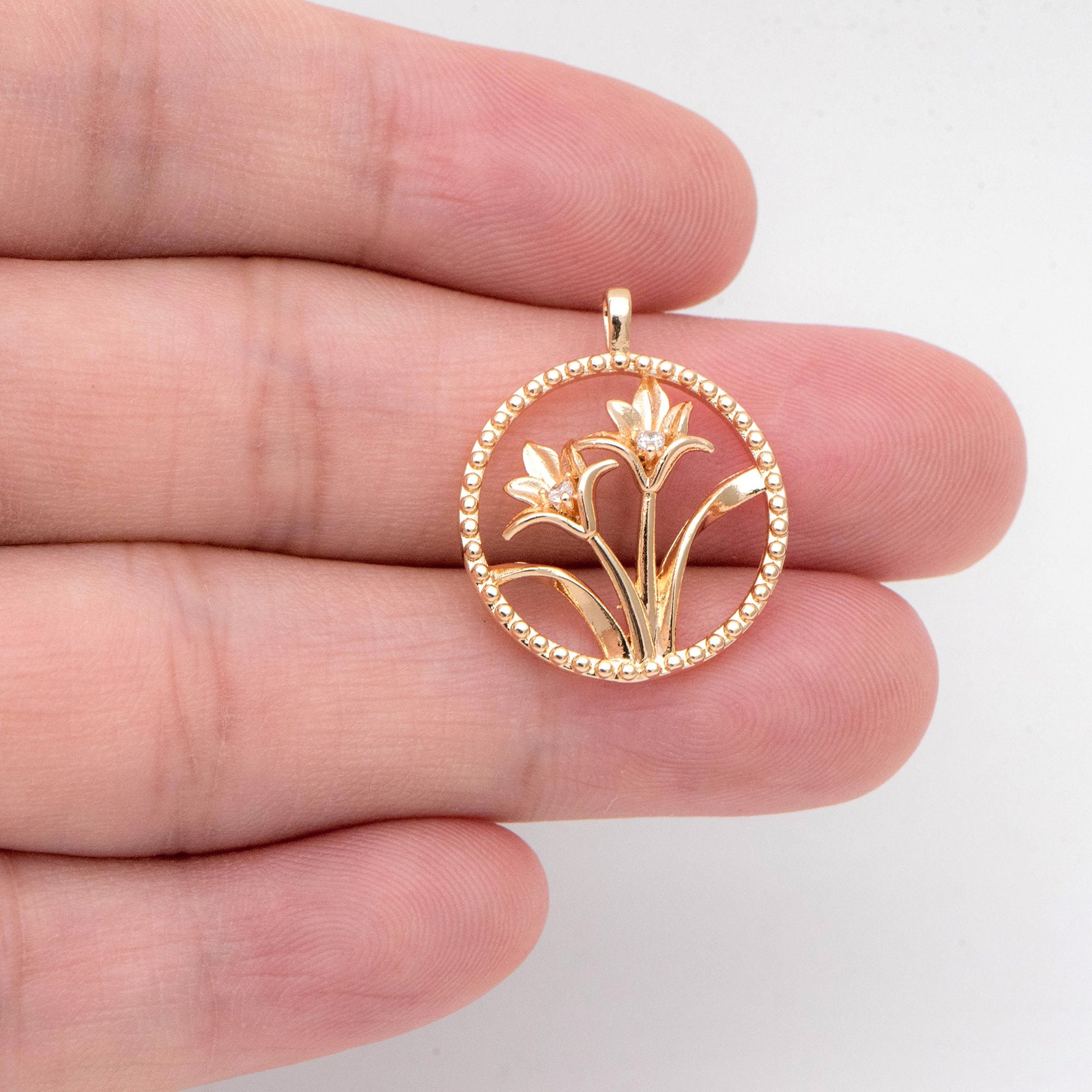 May include: Gold-colored pendant featuring a floral design within a beaded circle. The pendant showcases two stylized flowers with small, sparkling accents. The design is delicate and detailed, suitable for jewelry.