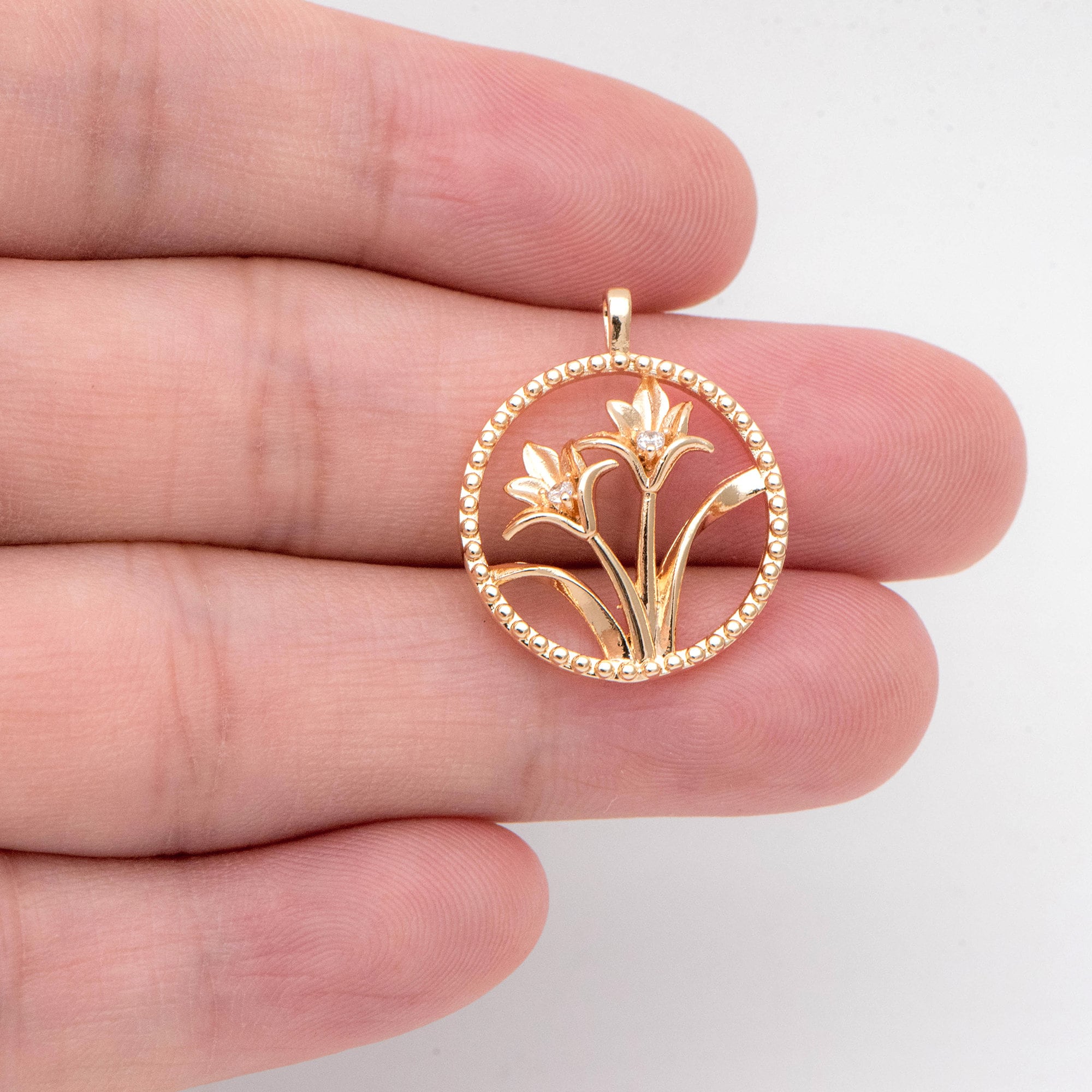 May include: Gold-colored pendant featuring a floral design within a beaded circle. The pendant showcases two stylized flowers with small, sparkling accents. The design is delicate and detailed, suitable for jewelry.