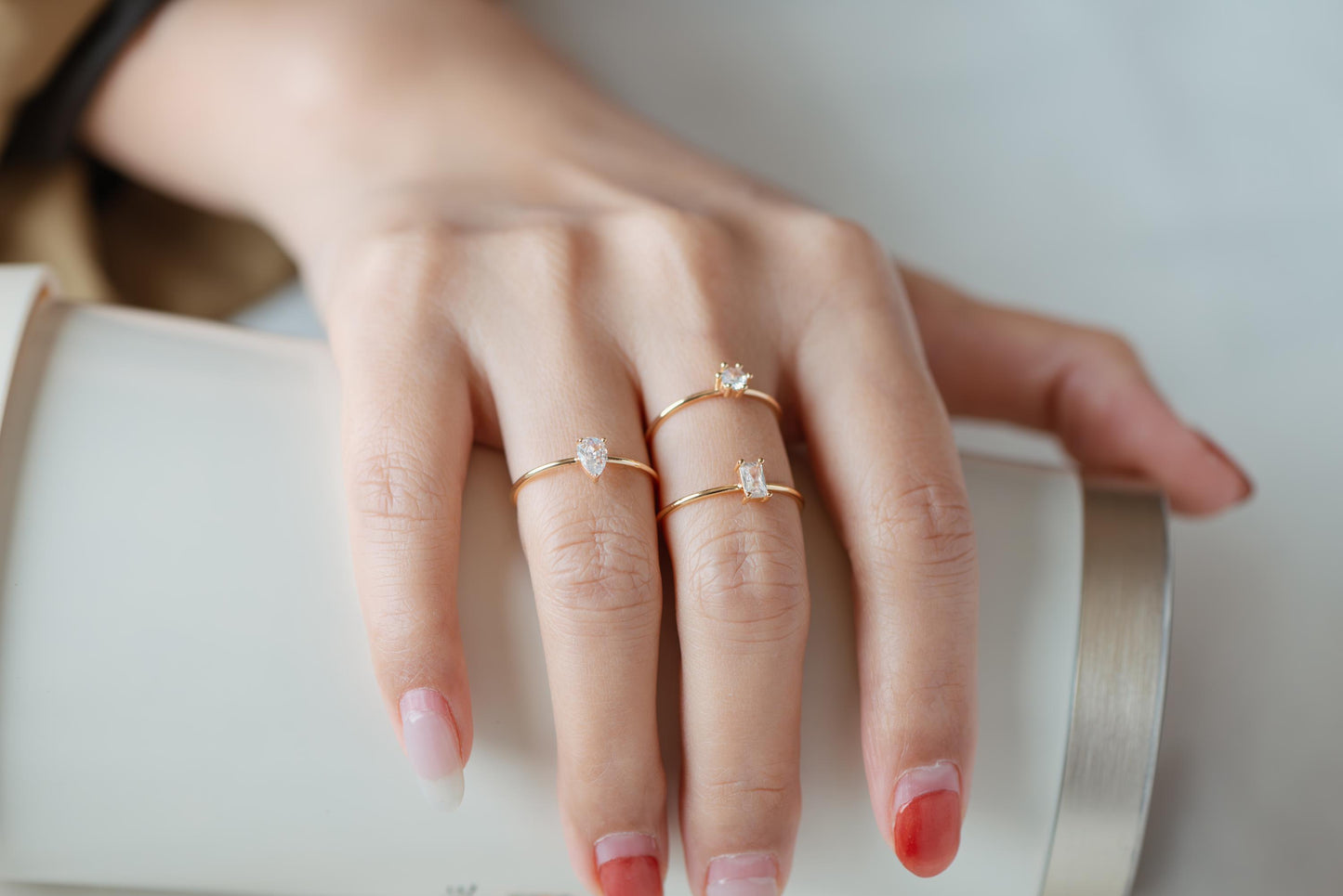 May include: Three delicate gold rings with clear gemstones are displayed on a hand. The rings feature different gemstone shapes: a pear, a square, and a round cut. The rings are set against a neutral background.