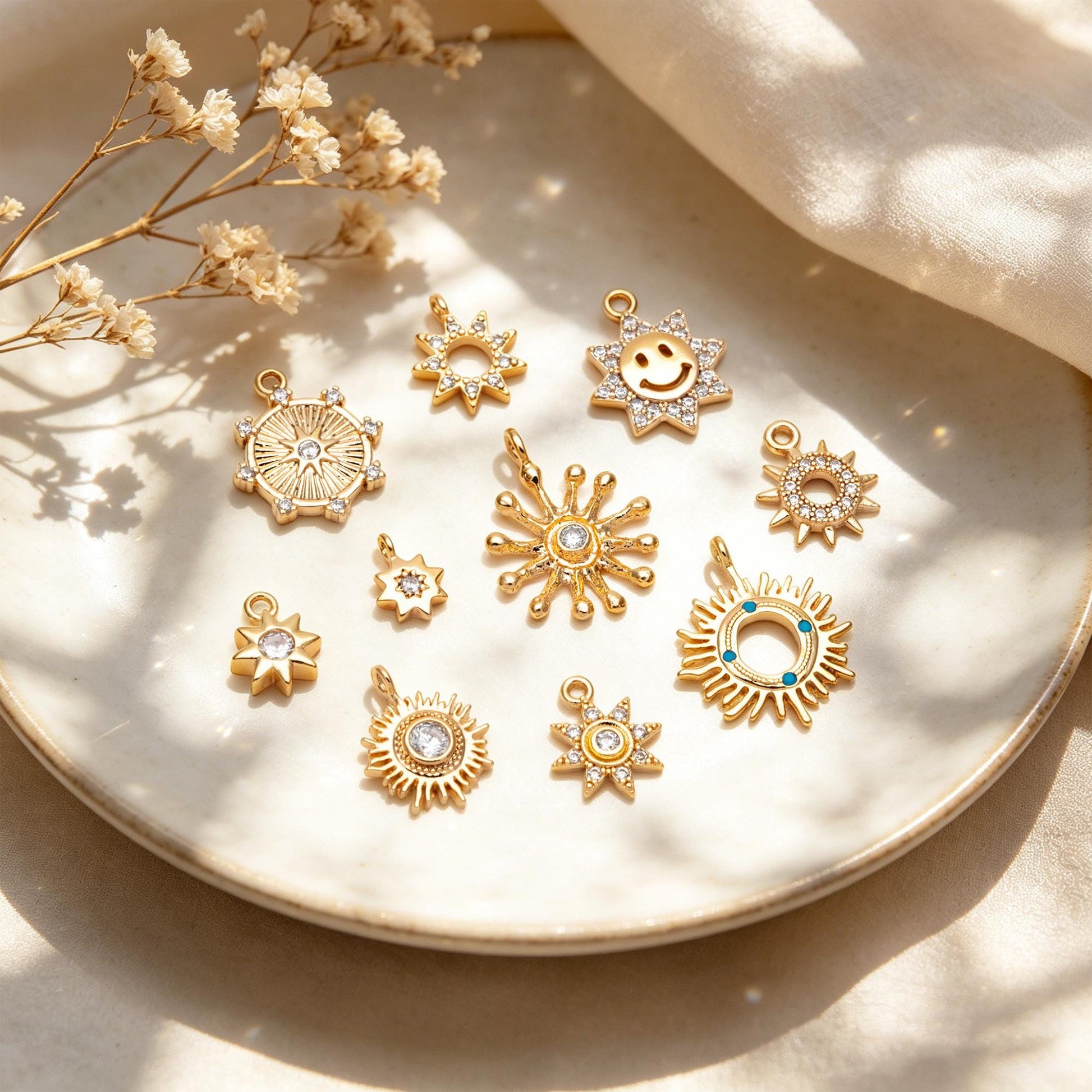May include: A collection of gold-colored sun-shaped charms with various designs, including a smiling face, starburst patterns, and embedded crystals. The charms are displayed on a white plate, suggesting jewelry or craft supplies.
