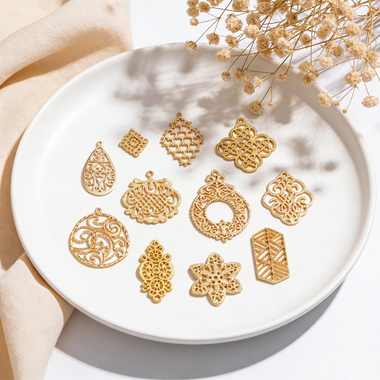 May include: A collection of gold-colored filigree charms in various shapes, including teardrops, squares, and floral designs, arranged on a white plate. The charms have intricate openwork patterns and are displayed with dried flowers.