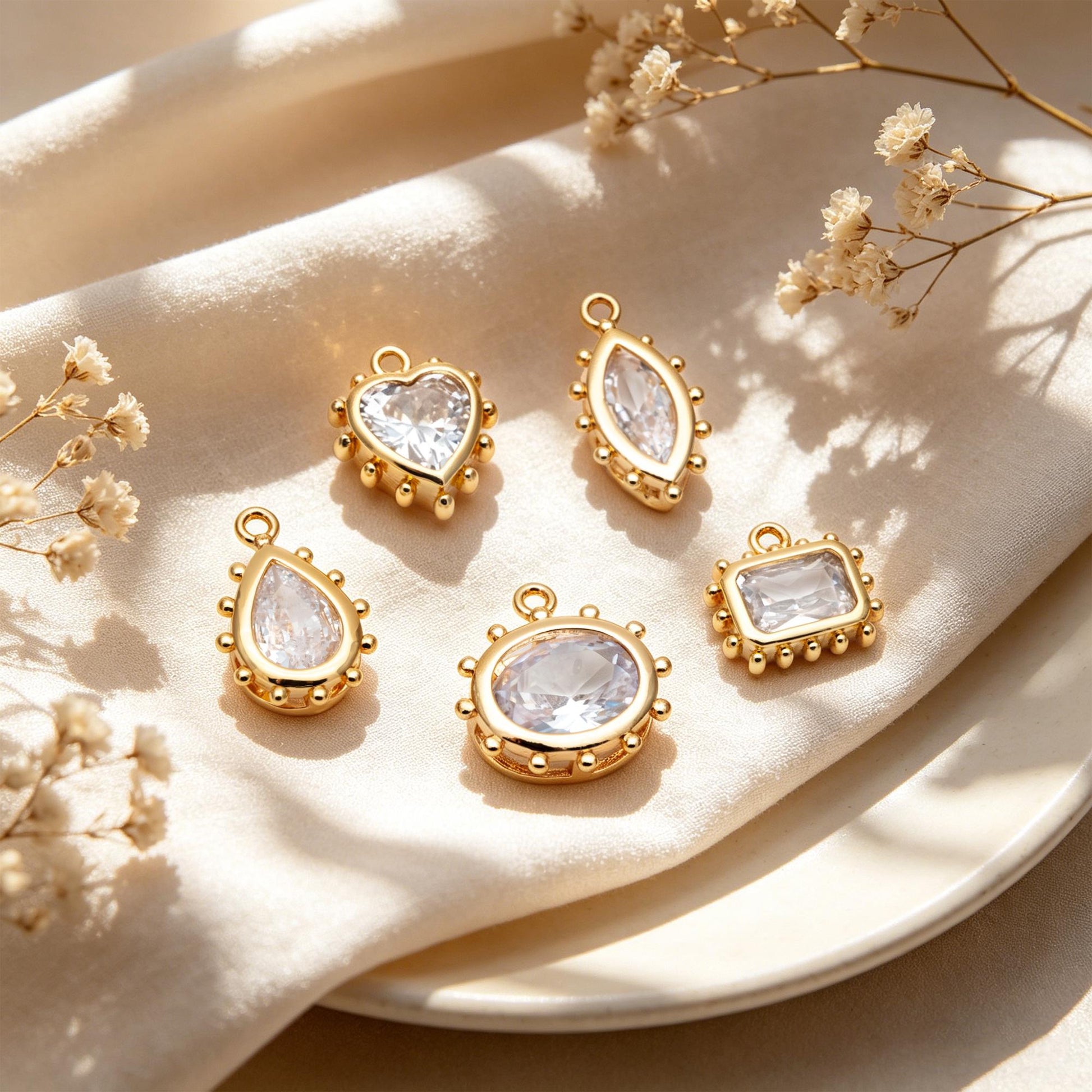 May include: Five gold-colored pendants with clear, faceted stones in various shapes: heart, marquise, teardrop, oval, and rectangle. Each pendant has a small loop at the top for hanging and is set against a cream-colored background.