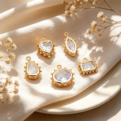 May include: Five gold-colored pendants with clear, faceted stones in various shapes: heart, marquise, teardrop, oval, and rectangle. Each pendant has a small loop at the top for hanging and is set against a cream-colored background.