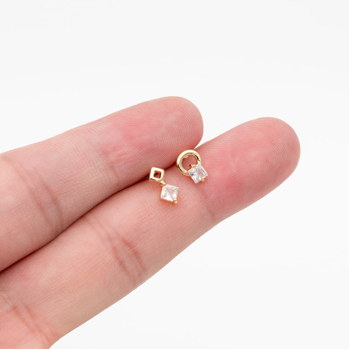 May include: A pair of gold earrings with a square-shaped clear stone and a small circle. The earrings are on a finger.