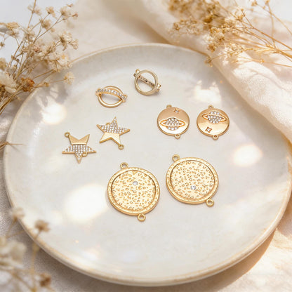 May include: A collection of gold-colored jewelry charms and rings, including star and planet designs, arranged on a white plate. Some charms are embellished with clear crystals, adding sparkle to the celestial-themed accessories.