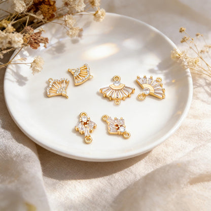 May include: Gold-toned jewelry charms in various fan and geometric shapes, adorned with clear crystals. The charms are displayed on a white plate, with dried flowers in the background. These charms are ideal for jewelry making.