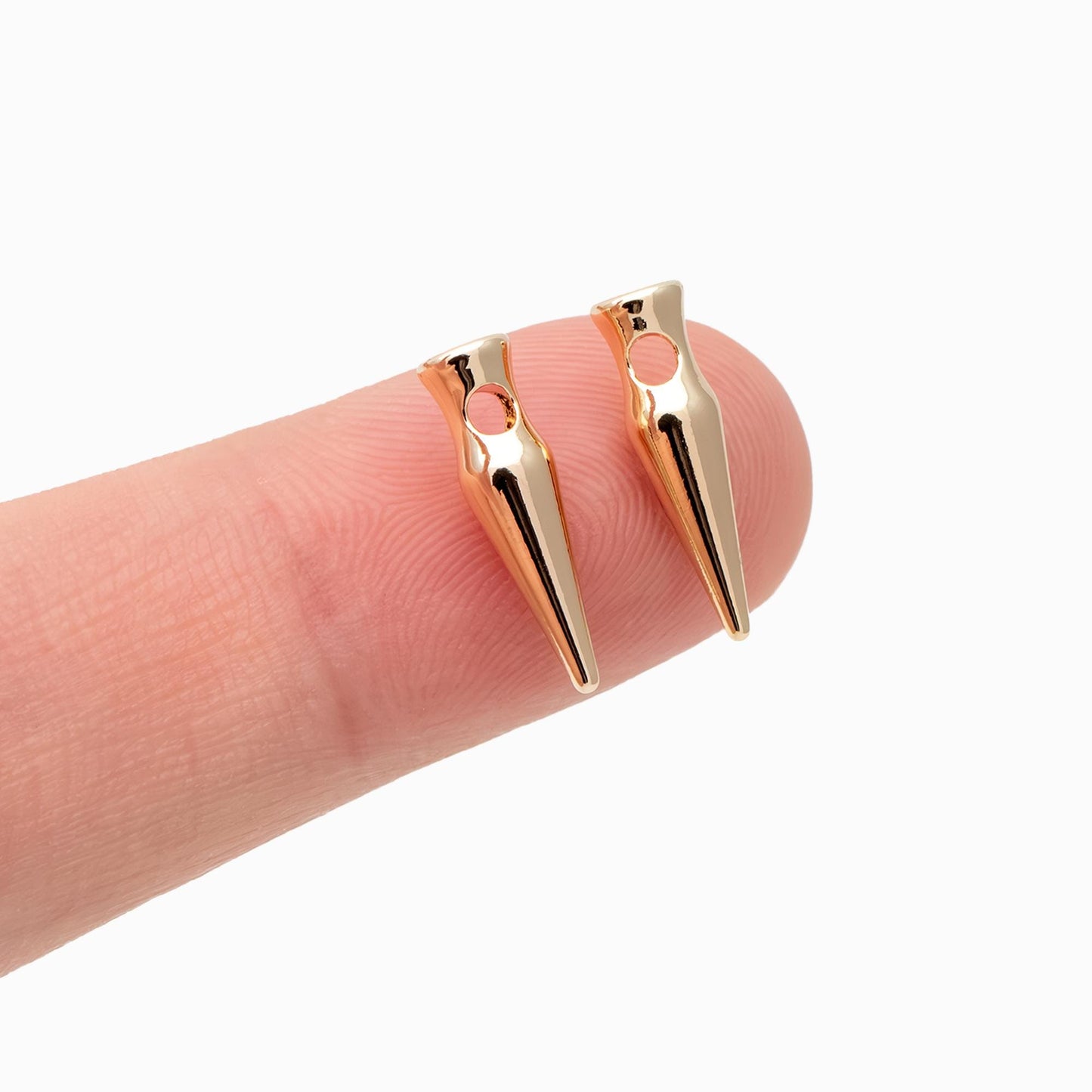 May include: Two gold-colored, cone-shaped jewelry findings with a small hole at the top. The findings are held on a finger against a white background. The findings have a polished, reflective surface.