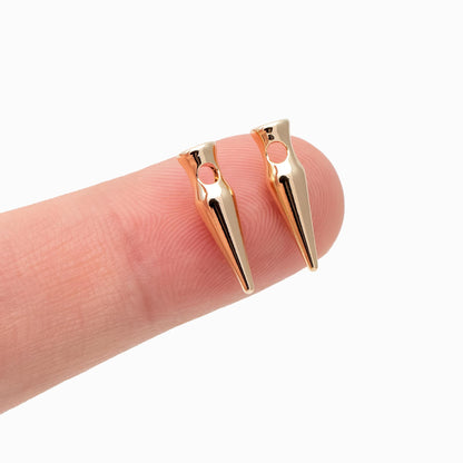 May include: Two gold-colored, cone-shaped jewelry findings with a small hole at the top. The findings are held on a finger against a white background. The findings have a polished, reflective surface.