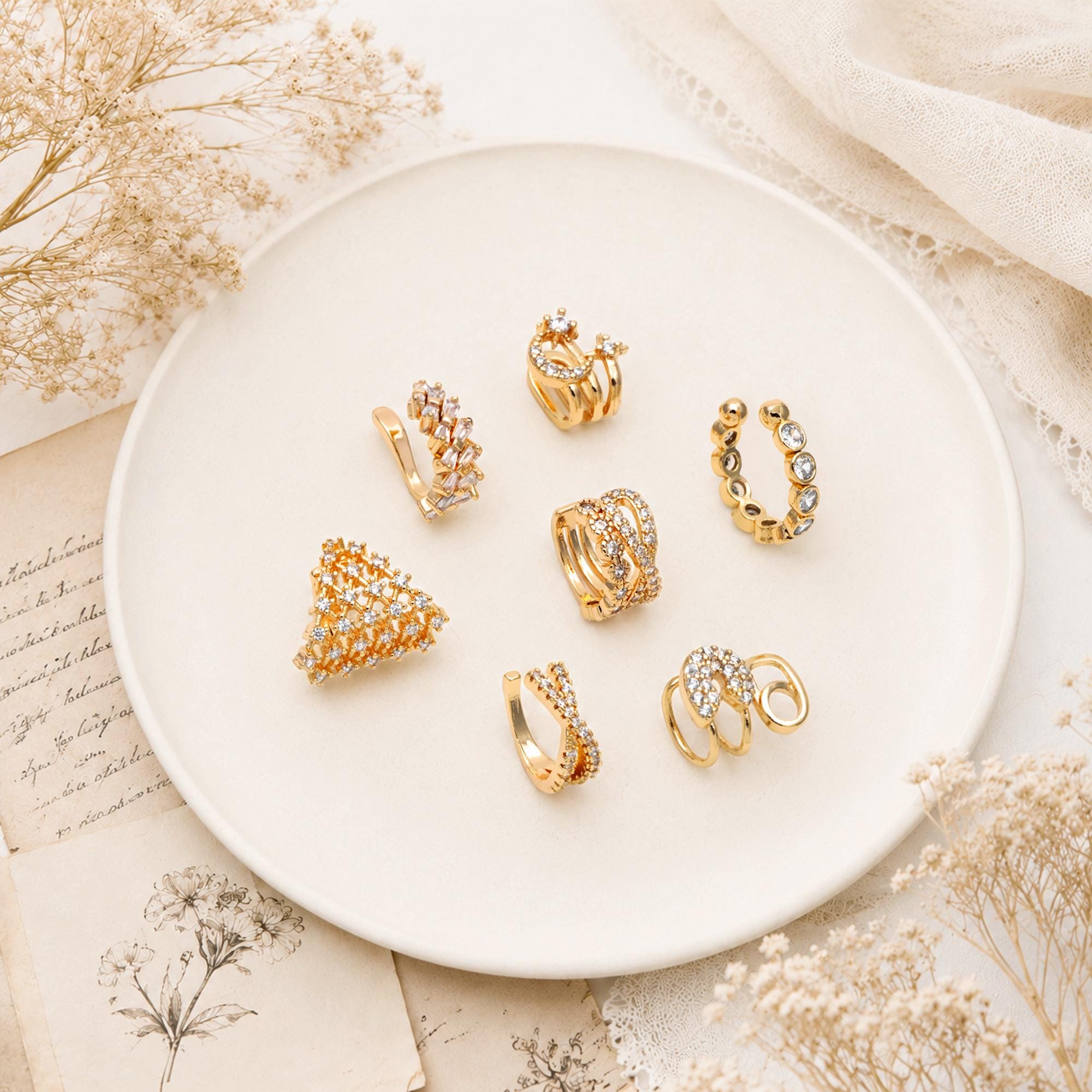 May include: A collection of gold-toned ear cuffs and earrings, adorned with clear crystals, arranged on a white plate. The jewelry pieces feature various designs, including geometric shapes and delicate curves, showcasing a range of styles for ear adornment.