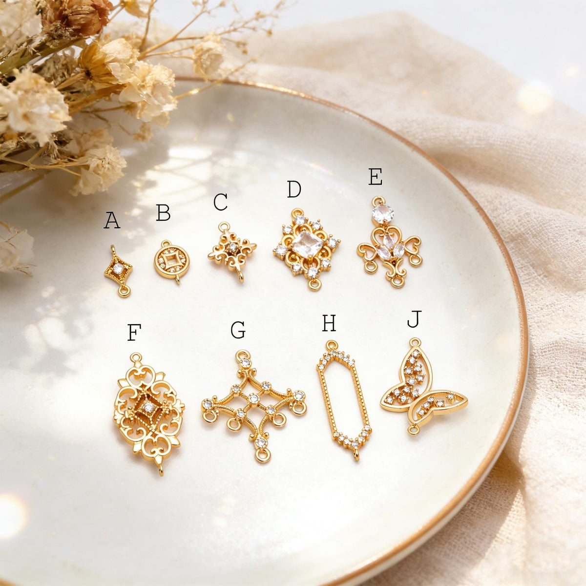 May include: A collection of gold-colored jewelry charms in various shapes, including diamonds, circles, crosses, and a butterfly. Some charms feature clear gemstones. The charms are displayed on a white plate.