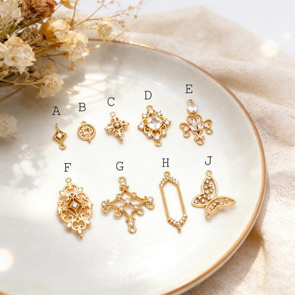 May include: A collection of gold-colored jewelry charms in various shapes, including diamonds, circles, crosses, and a butterfly. Some charms feature clear gemstones. The charms are displayed on a white plate.