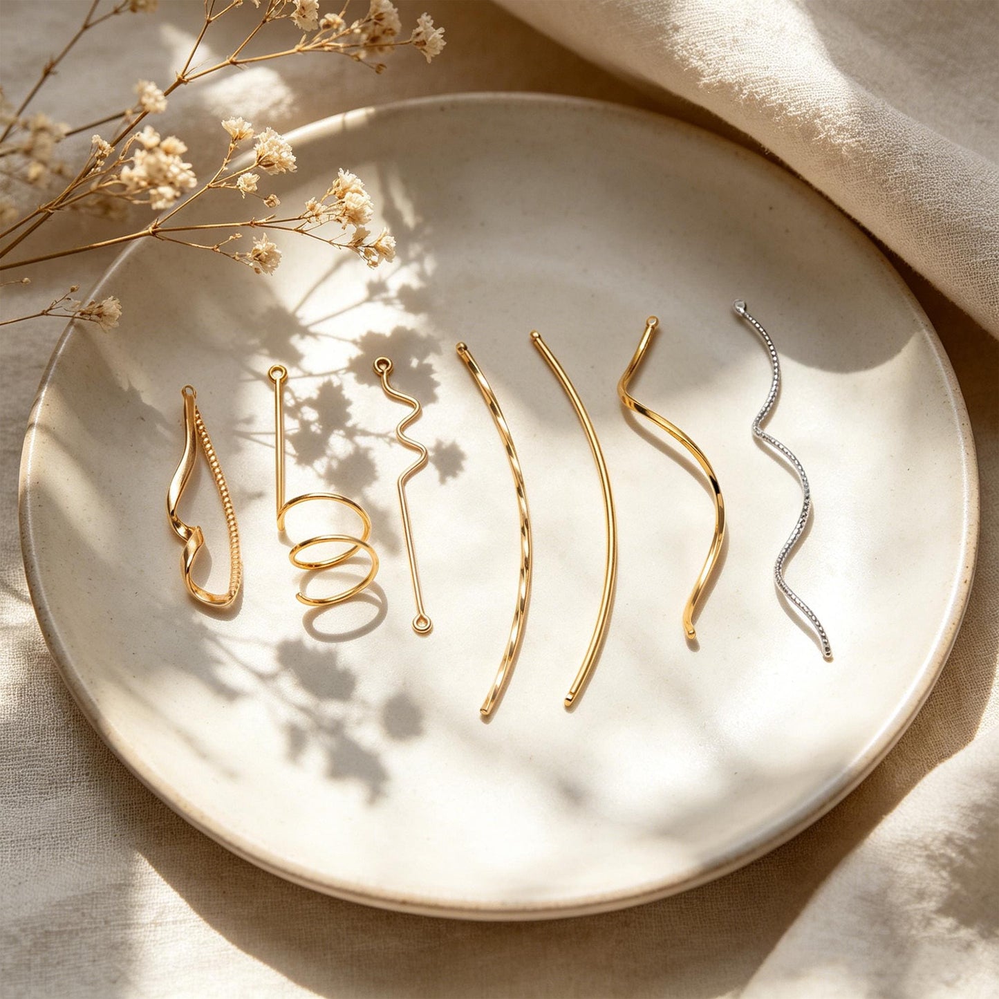 May include: A collection of gold and silver hair accessories, including barrettes and hair pins, arranged on a white ceramic plate. The accessories feature various designs, such as spirals, curves, and geometric shapes. Dried flowers are in the background.
