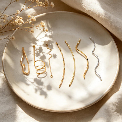 May include: A collection of gold and silver hair accessories, including barrettes and hair pins, arranged on a white ceramic plate. The accessories feature various designs, such as spirals, curves, and geometric shapes. Dried flowers are in the background.