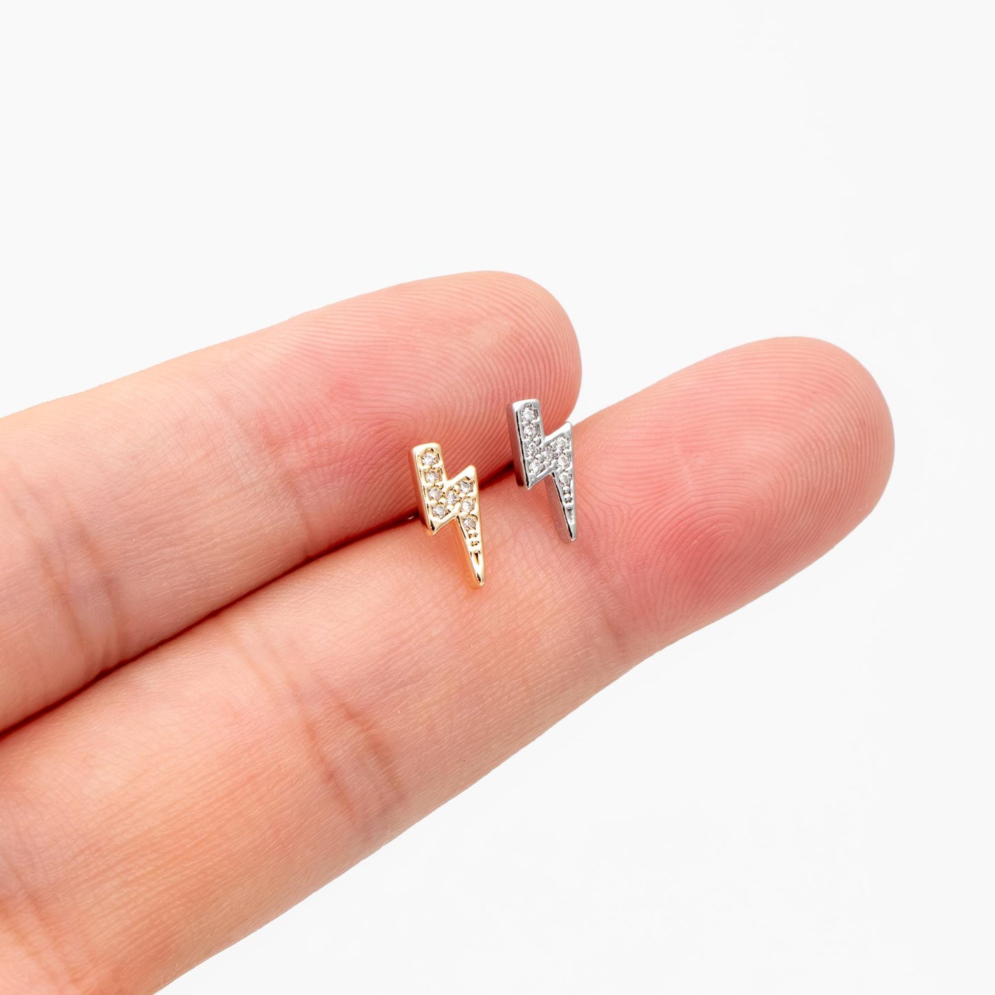 May include: Two small, stud earrings in the shape of lightning bolts. One is gold-toned, the other silver-toned, both encrusted with small, clear crystals. The earrings are displayed on a finger against a white background.