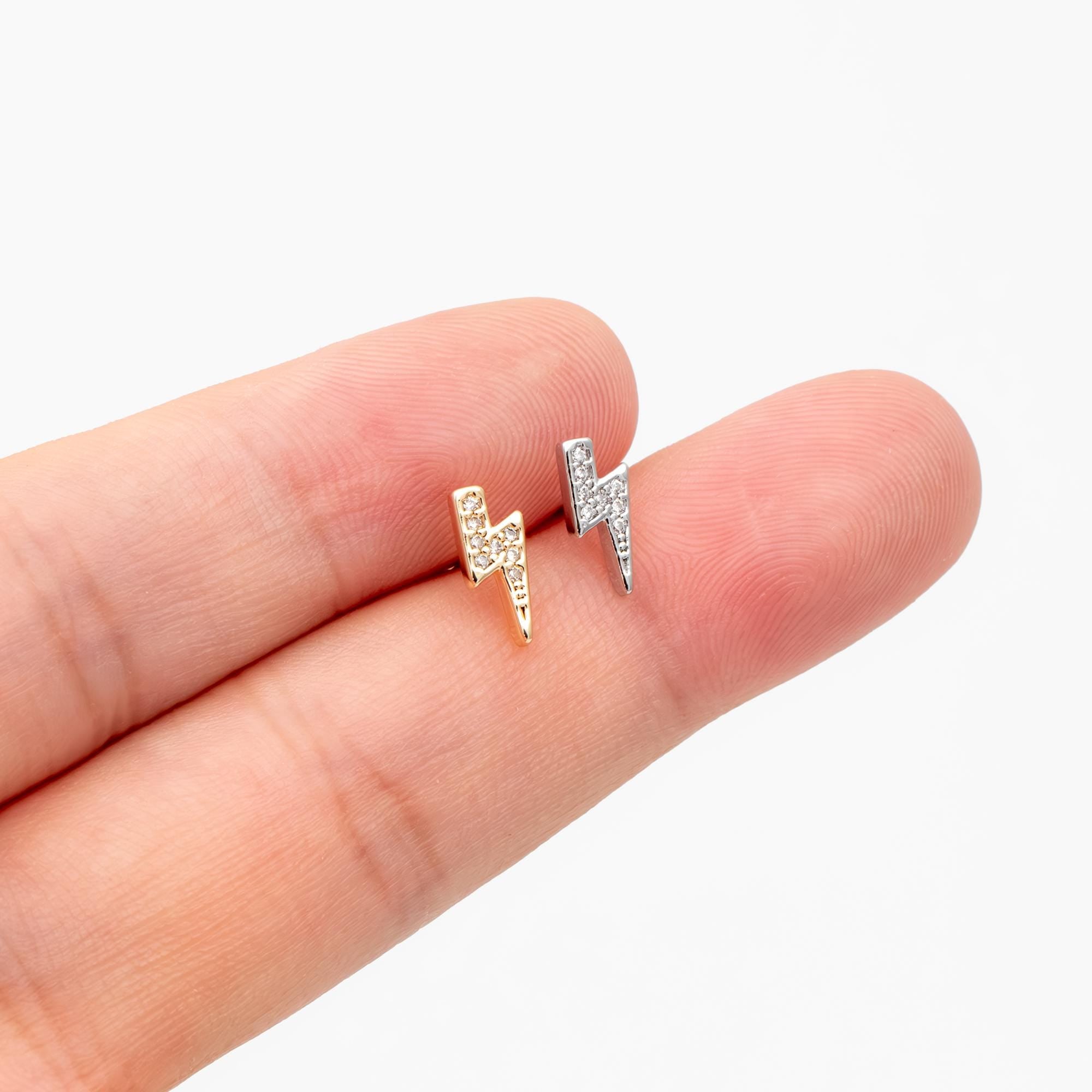May include: Two small, stud earrings in the shape of lightning bolts. One is gold-toned, the other silver-toned, both encrusted with small, clear crystals. The earrings are displayed on a finger against a white background.
