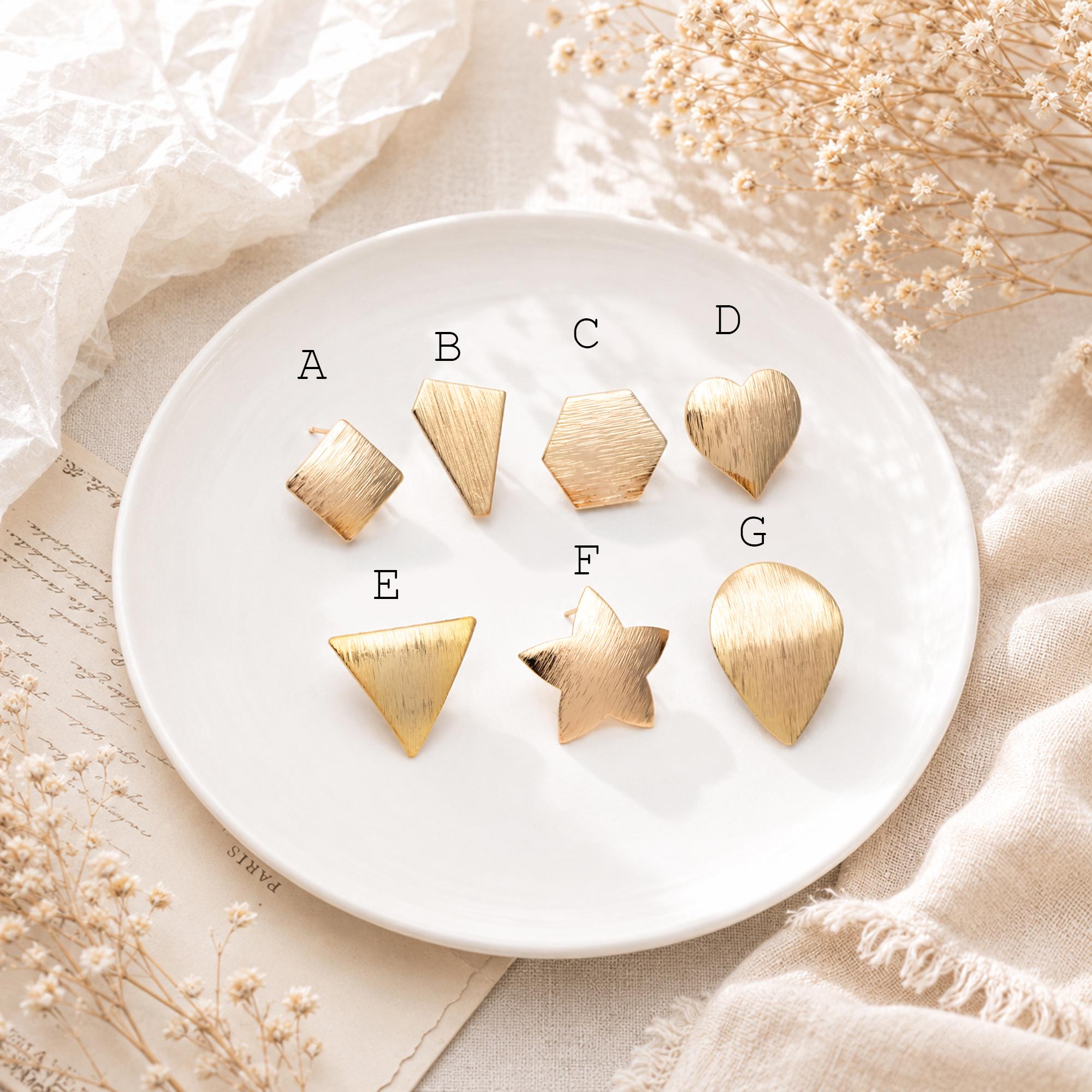 May include: A collection of seven gold-colored metal stud earrings in various geometric shapes, including a square, a trapezoid, a hexagon, a heart, a triangle, a star, and a teardrop. The earrings are arranged on a white plate.