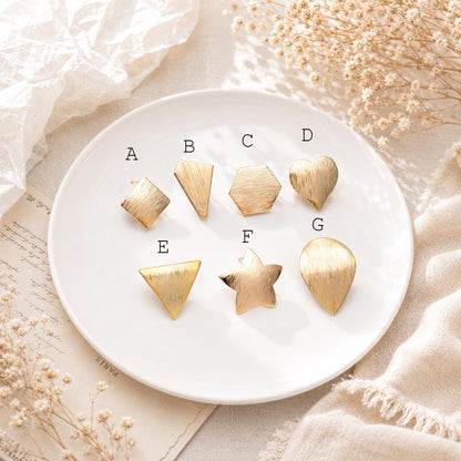 May include: A collection of seven gold-colored metal stud earrings in various geometric shapes, including a square, a trapezoid, a hexagon, a heart, a triangle, a star, and a teardrop. The earrings are arranged on a white plate.