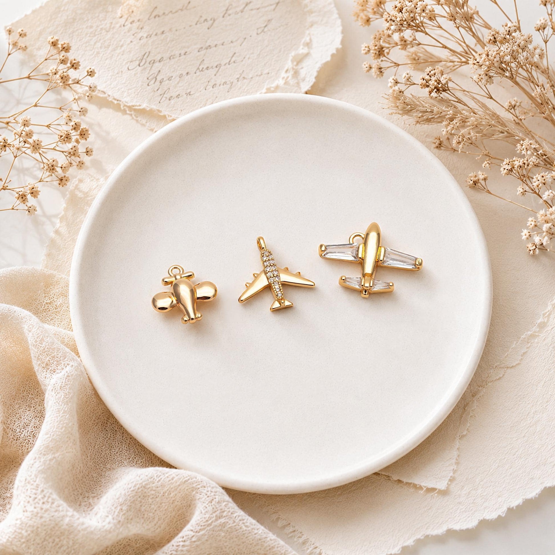 May include: Three gold-toned metal airplane charms arranged on a white plate. The first charm is a small rounded plane, the second is a slender plane with clear pavé stones along the fuselage, and the third is a larger plane with clear baguette stones.