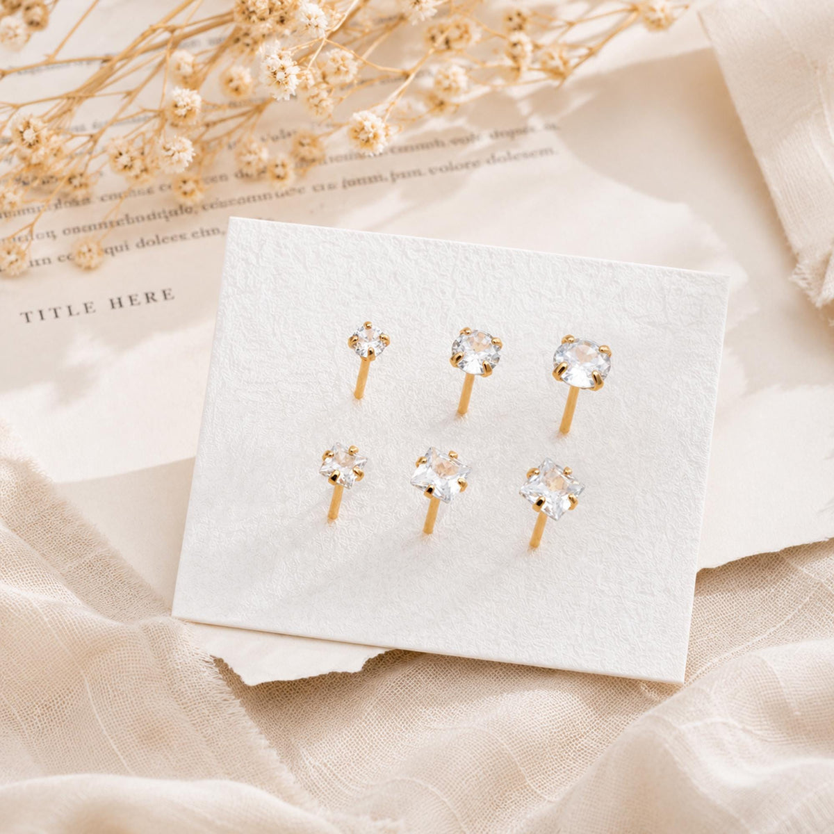 May include: A set of six gold-tone stud earrings arranged on a white display card. The earrings feature clear, faceted stones in various shapes, including round and square cuts, each secured with a gold-tone post and backing mechanism.