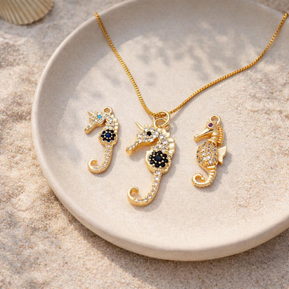 May include: A gold-colored seahorse pendant necklace and two matching seahorse charms. The seahorses are encrusted with clear and dark blue or red gemstones. The necklace and charms are displayed on a white plate, with a sandy background.