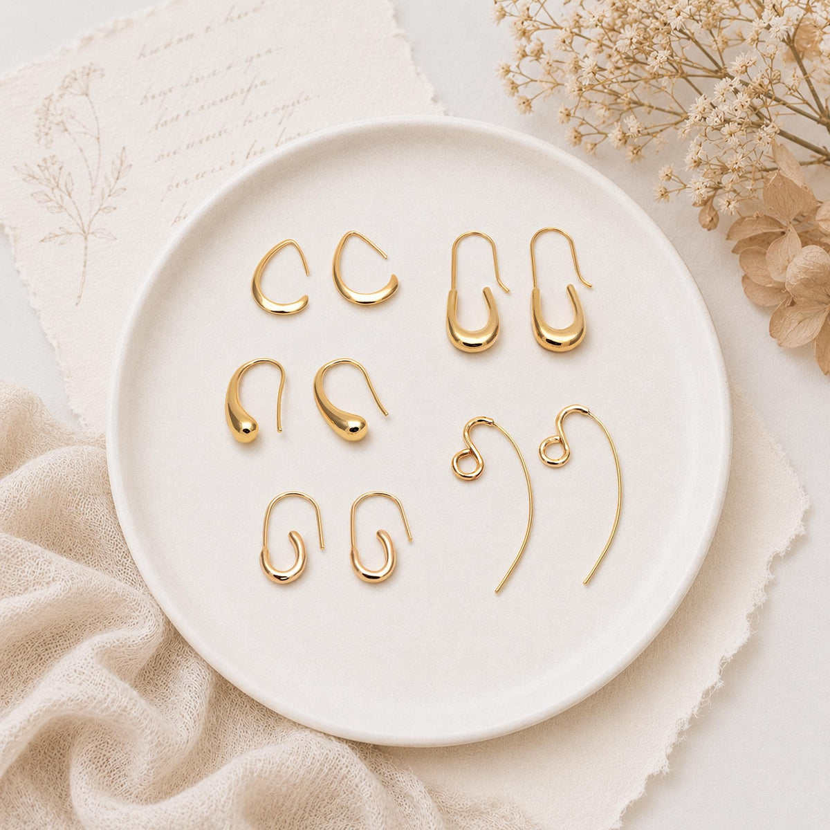 May include: Four pairs of gold-toned earrings arranged on a white plate. The collection includes small teardrop-shaped hoops, elongated U-shaped drop earrings, and a pair of curved wire earrings with a circular loop detail at the top of each piece.
