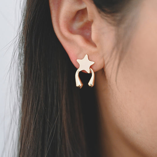 May include: Gold star-shaped earrings with a curved design. The earrings are worn in a person's ear.