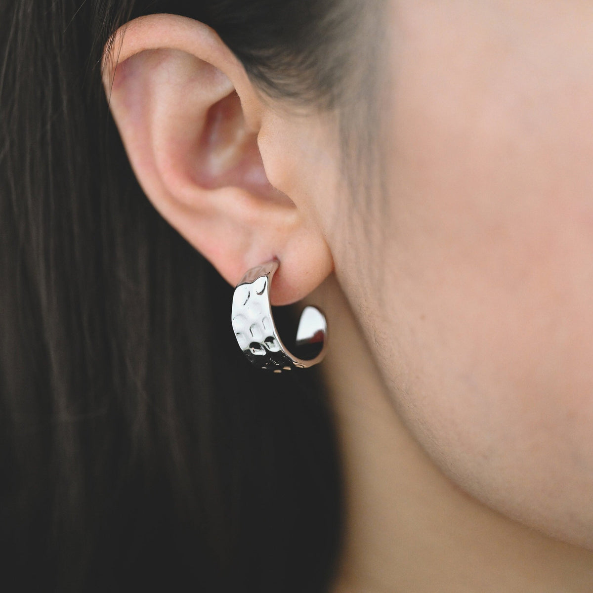 May include: Silver hoop earrings with a hammered texture. The earrings are worn in a person's ear.