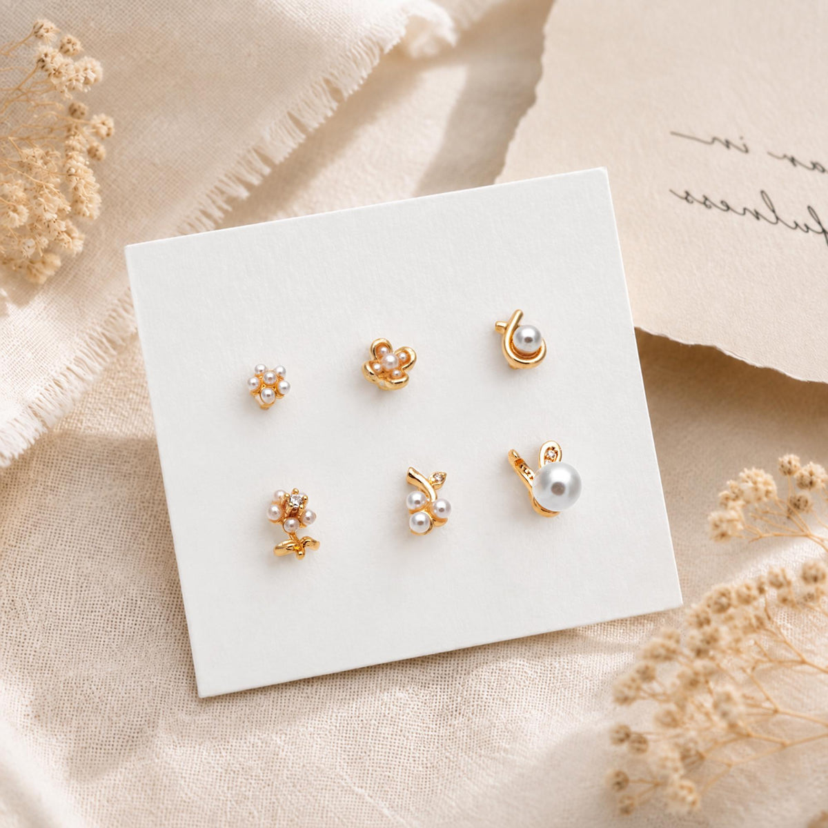 May include: A set of six gold-toned stud earrings displayed on a white card. The earrings feature various designs including small clusters of white pearls, floral-inspired shapes, and single pearl accents, all crafted with a polished gold-tone finish.