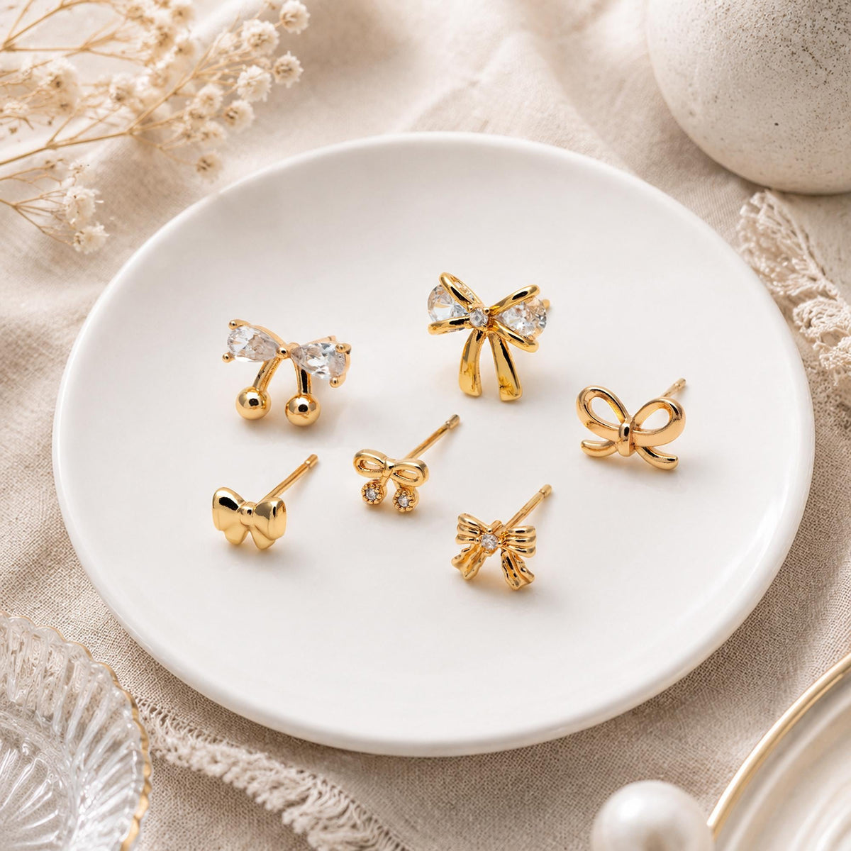 May include: A collection of six gold-tone stud earrings shaped like bows, arranged on a white plate. The designs include plain gold bows, bows accented with clear faceted crystals, and bow shapes featuring dangling crystal spheres.