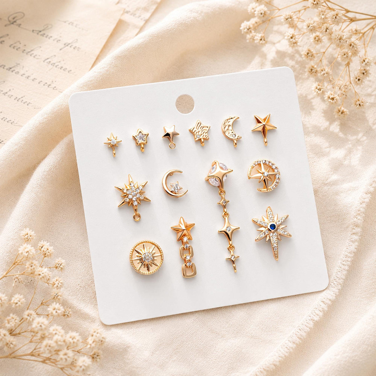 May include: A collection of gold-toned metal jewelry charms arranged on a white display card. The set includes various celestial-themed shapes such as stars, crescent moons, and sunbursts, some featuring clear crystals or a single blue stone accent.